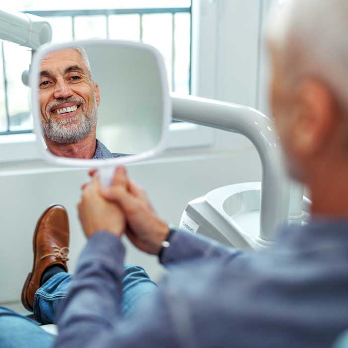 Cheerful senior man looking his teeth at the mirror while sitting at dentist's office